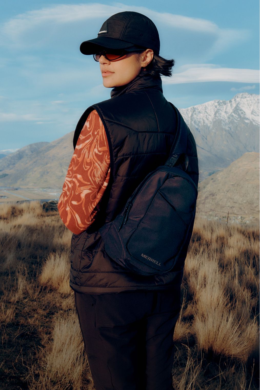 Woman wearing a Merrell vest and cap in a mountain landscape.